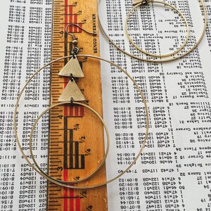 Peut inclure: Une paire de boucles d'oreilles cr&eacute;oles dor&eacute;es avec un pendentif triangulaire. Le pendentif est en m&eacute;tal et a une texture martel&eacute;e. Les boucles d'oreilles sont pr&eacute;sent&eacute;es sur une r&egrave;gle et un morceau de papier avec du texte.