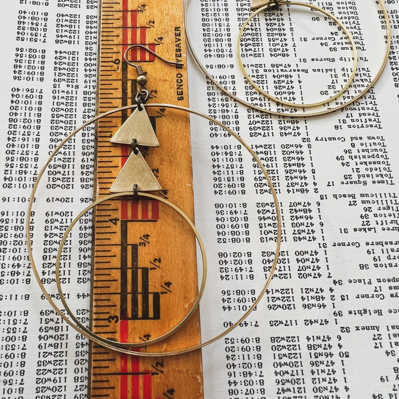 Peut inclure: Une paire de boucles d'oreilles cr&eacute;oles dor&eacute;es avec un pendentif triangulaire. Le pendentif est en m&eacute;tal et a une texture martel&eacute;e. Les boucles d'oreilles sont pr&eacute;sent&eacute;es sur une r&egrave;gle et un morceau de papier avec du texte.