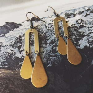 May include: A pair of gold-toned earrings with a geometric design. Each earring features a teardrop-shaped pendant and a curved arch.
