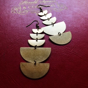 Puede incluir: Un par de pendientes de color dorado con un diseño en cascada de formas de semicírculo. Los pendientes están suspendidos de un pequeño gancho.