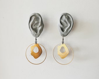 May include: A pair of earrings with a unique design. Each earring features a gray, textured, ear-shaped pendant with a gold, circular, layered design hanging from it.