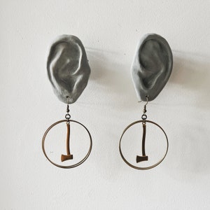 May include: A pair of earrings with a gray ear-shaped design and a gold hoop with a small axe charm hanging from each.