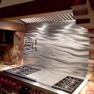 May include: A stainless steel backsplash with a wavy pattern, installed behind a gas stove in a kitchen. The backsplash is illuminated by overhead lights. The stove has a black cast iron grate and a stainless steel surface.