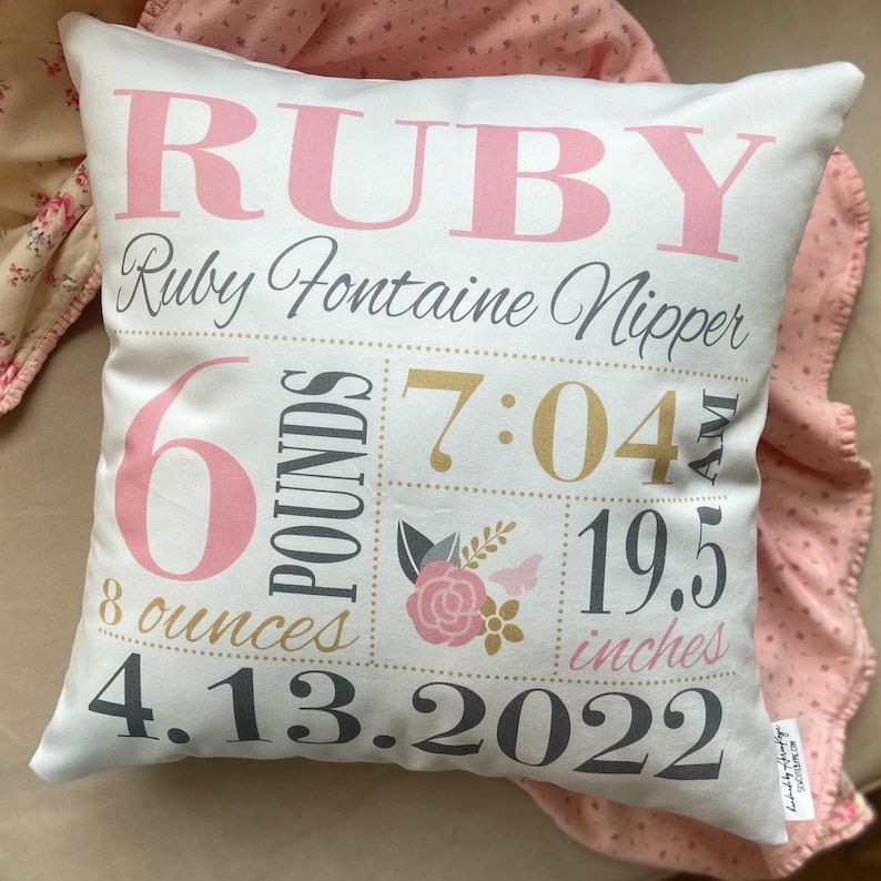 May include: A white pillow with pink and gold lettering. The pillow reads "RUBY", "Ruby Fontaine Nipper", "6 pounds 8 ounces", "7:04 AM", "19.5 inches", and "4.13.2022".