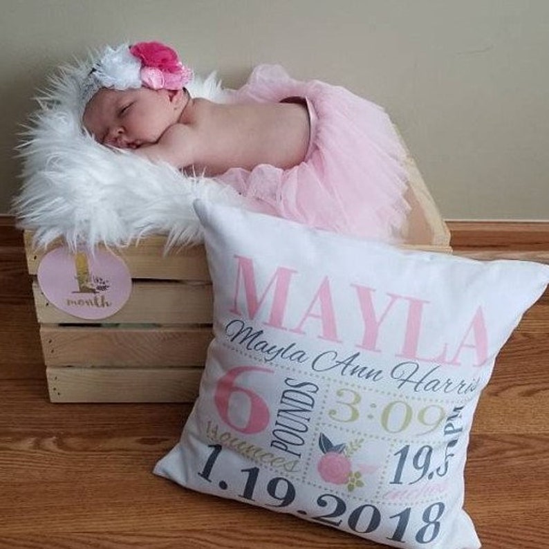 May include: A white pillow with pink and gray text that reads "Mayla Mayla Ann Harris 6 pounds 14 ounces 3:09 1.19.2018 19.5 inches". The pillow is decorated with pink flowers and green leaves. A baby girl is sleeping on a white fur rug in the background.