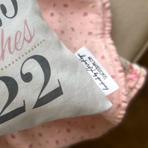 May include: White pillow with the text "Two Wishes 22" in black and pink lettering. The pillow has a small white tag with the text "Found by Ashley" and the website "SEW CUTE BY ME.COM".
