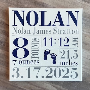 May include: A white canvas print with blue and gray text. The text reads "Nolan", "Nolan James Stratton", "8 pounds 7 ounces", "11:12 AM", "21.5 inches", and "3.17.2025".