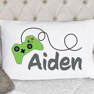 May include: A white rectangular pillowcase featuring a graphic of a light green video game controller with a black cord that loops around. The name Aiden is printed in large, dark gray, sans-serif letters below the controller graphic.
