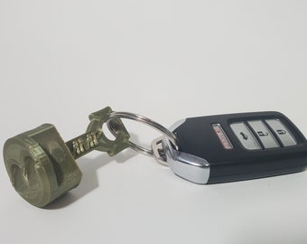 May include: A key ring with a 3D-printed green piston charm and a black car key fob. The key fob has silver accents and three buttons. The piston charm has the word "AMK" printed on it.