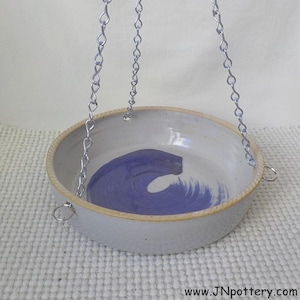 May include: A handmade ceramic hanging planter with a blue wave design inside. The planter is white with a tan rim and is suspended by silver chains. The website address www.JNpottery.com is visible at the bottom.