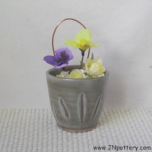 May include: A gray ceramic vase with a copper handle, holding yellow and purple flowers. The vase has a carved design and sits on a white, textured surface. The website address www.JNpottery.com is visible at the bottom.