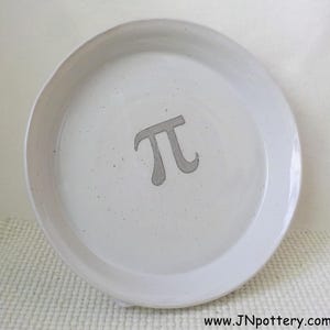 May include: A round, off-white ceramic pie plate with a gray Pi symbol in the center. The plate has a slightly raised edge and a speckled texture. The website address www.JNpottery.com is visible at the bottom.