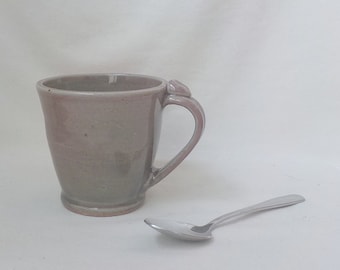 May include: A gray ceramic mug with a handle and a slight glaze variation. The mug is sitting on a white surface next to a silver spoon.