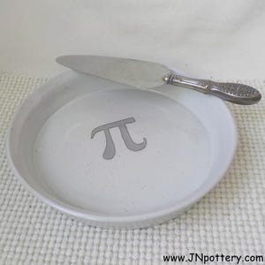 May include: A white ceramic pie plate with a gray "pi" symbol in the center. A silver pie server rests on the plate. The plate has a slightly speckled texture and a simple, elegant design. The pie server has a decorative handle.