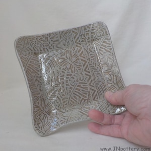 May include: Square ceramic dish with a leaf pattern in shades of beige and white. The edges are slightly curved. The dish is held by a hand.