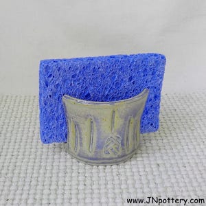 May include: A ceramic sponge holder with a light purple and yellow glaze featuring vertical carved lines and a central knot design. A rectangular blue textured sponge is positioned upright inside the holder. The text www.JNpottery.com is visible.
