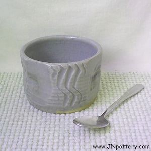 May include: A small, light gray ceramic bowl with a wavy, textured design. The bowl is sitting on a white, textured surface. A silver spoon is lying next to the bowl.
