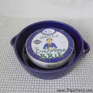 May include: A round of Petit Bonhomme Brie cheese, labeled "Grass Fed," sits inside a deep blue ceramic dish with handles. The cheese packaging features a cartoon illustration of a man. The dish has a textured rim.