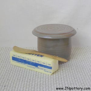 May include: A ceramic butter dish with a lid, a wooden butter knife, and a stick of butter. The butter dish is a muted gray-brown color. The butter is in a yellow wrapper with blue text. The knife is light brown.