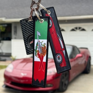 May include: A keychain with two keychains attached. One keychain has a red, white, and green design with a stylized eagle. The other keychain has a red car with black wheels on a black background. The keychains are hanging from a silver carabiner.