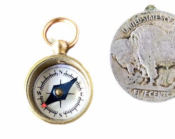 May include: A small brass compass with a white face and black needle. The compass has a gold loop at the top for attaching to a chain or lanyard. The compass is about the size of a US five-cent coin.