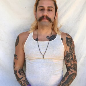 May include: A man with long blonde hair and a mustache is wearing a white tank top and has several tattoos on his arms. He is wearing a necklace with a pendant.