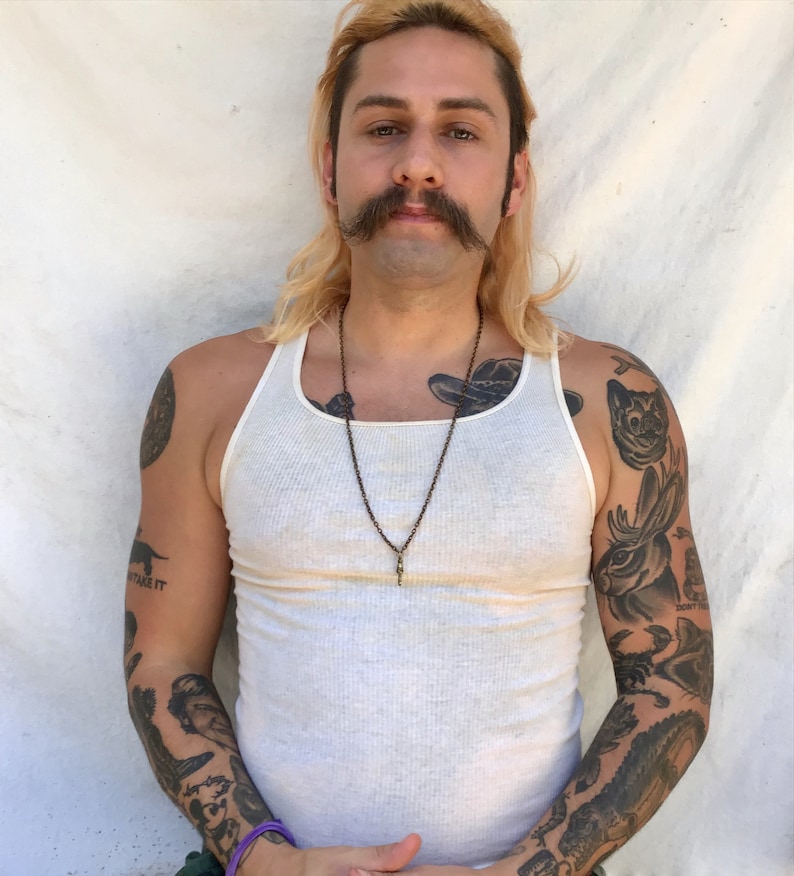 May include: A man with long blonde hair and a mustache is wearing a white tank top and has several tattoos on his arms. He is wearing a necklace with a pendant.