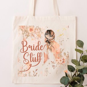 May include: A white canvas tote bag with a floral design and the text "Bride Stuff" in a copper color. The design features a watercolor illustration of a bride in a white dress with her hair pulled back.