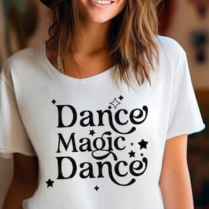 Pode incluir: Camiseta branca com as palavras "Dance Magic Dance" em preto, com uma fonte caprichosa, adornada com estrelas. A camiseta tem gola redonda e mangas curtas. O design é simples e elegante.