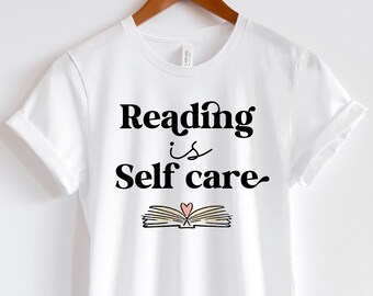 May include: White t-shirt with the text "Reading is Self care" in black script. Below the text is an illustration of an open book with a heart. The shirt is hanging on a wooden hanger.