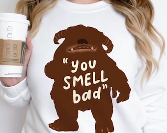 May include: White sweatshirt featuring a brown monster graphic with the text "you SMELL bad" in a playful font. The monster has horns and a toothy grin. A coffee cup with a brown sleeve is held in the background.