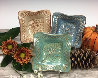 May include: Three square ceramic dishes with wavy edges, each featuring a central leaf impression and a border of small leaves. The dishes are glazed in cream, blue, and teal, with dark brown accents highlighting the textured leaf patterns.