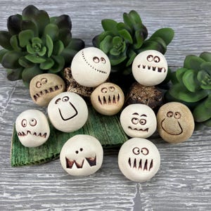 May include: Ten white ceramic spheres with different hand-painted faces. Each sphere has a unique expression, including eyes, mouths, and other facial features. The spheres are arranged on a green and brown surface.