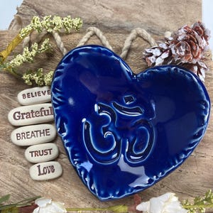 May include: A glossy, deep blue heart-shaped ceramic dish with the Om symbol embossed in the center. The dish has a rope hanger and is surrounded by small stones with inspirational words like "Believe" and "Love". Decorative sprigs and a pinecone add to the aesthetic.