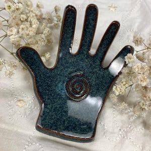 May include: A ceramic hand-shaped dish in shades of teal and brown. The dish features a spiral design in the center and a glossy finish. The edges are outlined in brown. White flowers are in the background.