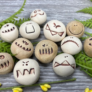 May include: Ten small, round, white ceramic stones with different brown painted faces. Each stone has a unique expression, including happy, sad, angry, and surprised.
