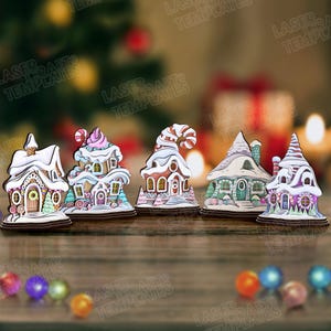 May include: A collection of five wooden Christmas village ornaments. Each ornament is a unique gingerbread house design with colorful details, including snow-covered roofs, candy accents, and decorative doors. The ornaments are set on a wooden surface.