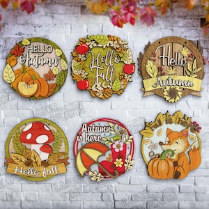 Könnte beinhalten: Sechs Holzschilder mit lasergeschnittenen Herbstmotiven. Jedes Schild zeigt herbstliche Elemente wie Kürbisse, Blätter, Äpfel und einen Fuchs. Der Text lautet "Hello Autumn", "Hello Fall" und "Autumn is here."