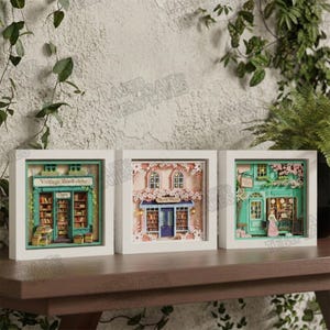 Op de afbeelding: Drie ingelijste papieren kunstwerken die miniatuur boekhandels uitbeelden. De eerste is een vintage boekhandel met groene randen en open deuren. De tweede heeft een blauwe deur en bloemmotieven. De derde toont een groene winkel.