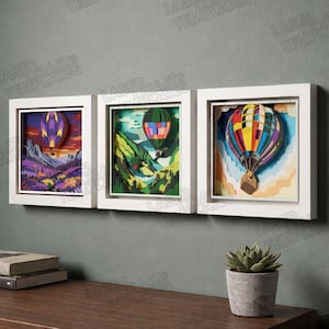 May include: Three framed 3D art pieces featuring hot air balloons. Each piece has a white frame and depicts a colorful balloon against a landscape. The first shows a purple and yellow balloon over a sunset. The second shows a green balloon over a mountain range. The third shows a multi-colored balloon.