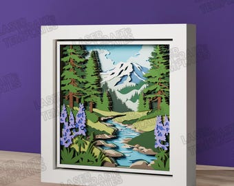 Könnte beinhalten: Ein mehrschichtiger Papierschattenkasten mit einer Berglandschaft, einem schneebedeckten Gipfel, immergrünen Bäumen, einem gewundenen blauen Fluss und violetten Lupinenblüten im Vordergrund, eingefasst in einen quadratischen weißen Rahmen.