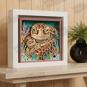 May include: A layered paper art piece featuring two sea turtles in a square white frame. The turtles are brown with intricate details, surrounded by colorful coral and aquatic plants. The artwork is displayed on a wooden surface.