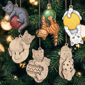 Pode incluir: Seis ornamentos pendentes com gatos a interagir com bolas redondas. Três são coloridos, mostrando um gato cinzento com uma bola vermelha, um gato laranja com uma bola verde e um gato branco com uma bola amarela. Três são de madeira natural gravada.