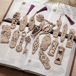 Könnte beinhalten: Eine Sammlung lasergeschnittener Lesezeichen aus Holz mit verschiedenen Fantasy-Motiven, darunter ein Schild, Kristall, Feder, Axt, Trankflasche, Schlüssel, Drache, Turm und Sanduhr, einige davon mit violetten Quasten, auf einem offenen Buch.