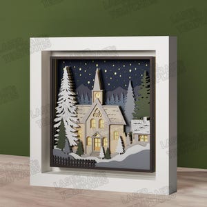 Puede incluir: Una obra de arte en caja de sombra multicapa que muestra una escena de iglesia invernal. El diseño incluye un edificio de iglesia central, pinos cubiertos de nieve y un cielo nocturno azul oscuro con estrellas amarillas, en un marco blanco.