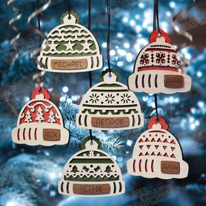 Puede incluir: Adornos navideños de madera con forma de gorros, cada uno con un diseño único y un nombre. Los adornos son blancos con detalles rojos y verdes, y los nombres incluyen Michael, Nick, George, Bonnie, Pettie y Damon.