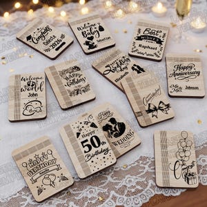 May include: A collection of wooden rectangular plaques with engraved messages for various occasions. Messages include "Congrats," "Welcome Baby," "Happy Birthday," and "Happy Anniversary." The plaques feature decorative borders and are arranged on a lace tablecloth.