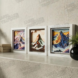 Peut inclure: Trois œuvres d'art encadrées représentant des montagnes. Chaque pièce présente un design en papier superposé d'une scène de montagne dans un cadre blanc. Les palettes de couleurs comprennent des nuances de violet, d'orange, de bleu et de marron. Les œuvres sont exposées sur une surface grise.