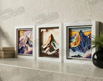 May include: Three framed mountain art pieces. Each piece features a layered paper design of a mountain scene in a white frame. The color palettes include shades of purple, orange, blue, and brown. The art pieces are displayed on a gray surface.