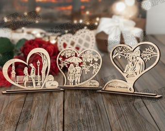 May include: Three wooden heart-shaped decorations. Each features a couple in a romantic scene, laser-cut from light-colored wood. One shows a couple by a lamppost, another under an umbrella, and the last under a tree. Names are engraved on each base.